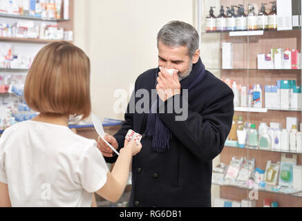 Sick man with flu consulting with pharmacist about medicaments. Female specialist in white coat offering blister pack of helpful pills. Man having blocked nose, blowing nose with tissue, coughing. Stock Photo