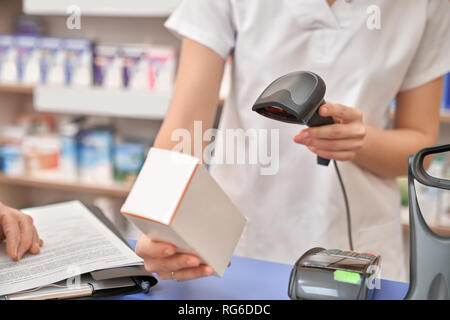 Cashier in drugstore scanning price using barcode scanner. Female pharmacist holding medical box with medicaments. Woman standing at counter, wearing in white uniform. Stock Photo