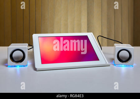 On the table is a tablet computer mounted on a tablet PC holder and connected to a two small white speaker with LED lighting. Stock Photo