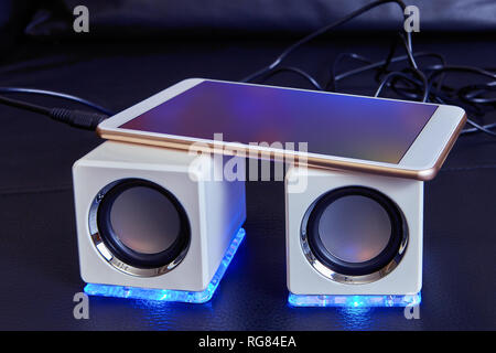 Two white mini speakers, of cubic form, with blue LED lights, are connected to the smartphone with a cable, the smartphone lies on top of the speakers Stock Photo