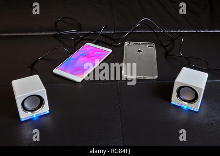 On sofa lies smartphone to which small white cubic-shaped speakers made of plastic and equipped with blue LED light are connected with cable, and sili Stock Photo