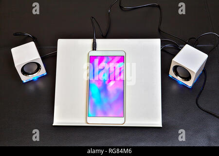 The smartphone is placed in a plastic holder and connected to white mini speakers with blue led backlight. Stock Photo