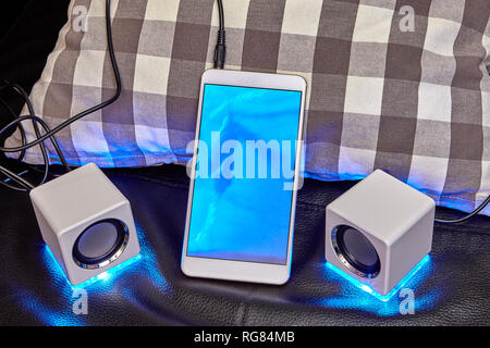 A smartphone with a blue display is connected to the mini speaker with a cable. Stock Photo
