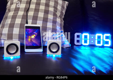 On the sofa there are led speakers and a clock with a blue luminous dial, speakers are connected to the smartphone with a cable. Stock Photo