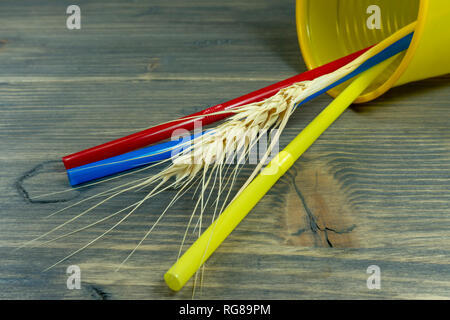 Three colored plastic straw lying on their sides in a yellow beaker with an ear of ripe golden wheat on wood Stock Photo