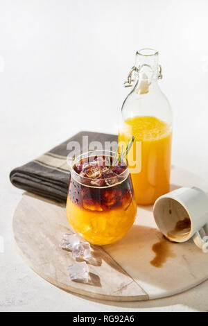 Ice cold summer drink. Orange juice espresso with ice, bottle of organic orange juice and empty espresso shot on white marble serving plate over white Stock Photo