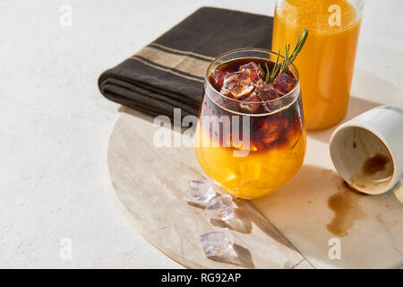 Ice cold summer drink. Orange juice espresso with ice, bottle of organic orange juice and empty espresso shot on white marble serving plate over white Stock Photo