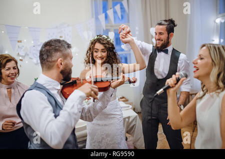 A young cheerful bride, groom and other guests dancing, singing and playing violin on a wedding reception. Stock Photo