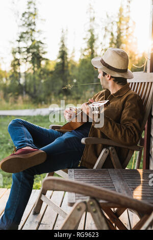 Young man sitting on veranda of a wood house, playing the ukulele Stock Photo