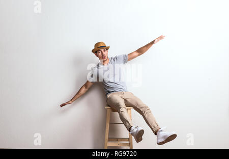 A confident hispanic young man with hat sitting on a stool in a studio, arms stretched. Stock Photo