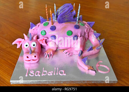 Home made cake for six years old childs birthday celebration with candles on cake board with table background featuring caricature of pink dragon Stock Photo