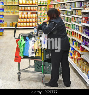 Woman picker working selecting products on supermarket shelves & place in plastic bags onto trolley for home food grocery delivery via van England UK Stock Photo