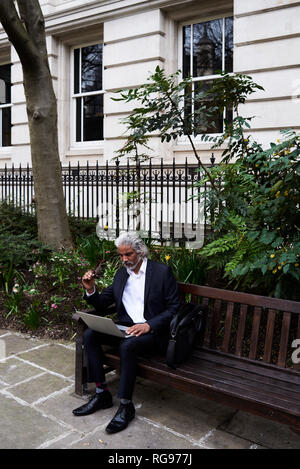Senior businessman sitting on bench outdoors working on laptop Stock Photo