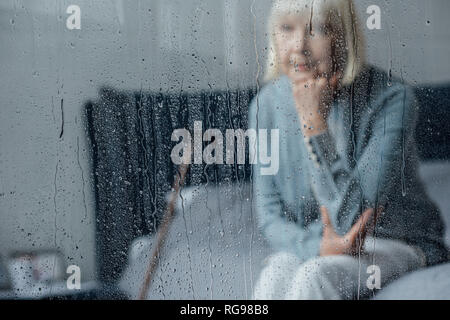 lonely senior woman sitting on bed and propping chin with hand at home through window with raindrops Stock Photo