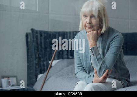 sad senior woman sitting on bed and propping chin with hand at home Stock Photo