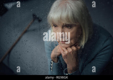 depressed senior woman sitting and propping chin with hands at home through window with raindrops Stock Photo