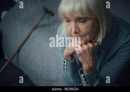 sad senior woman sitting and propping chin with hands at home through window with raindrops Stock Photo