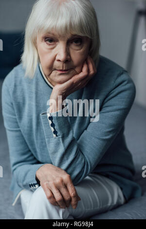 upset senior woman with grey hair propping chin with hand and looking at camera at home Stock Photo