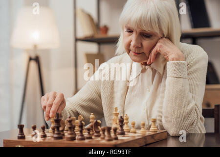 pensive senior woman sitting at table and playing chess at home Stock Photo