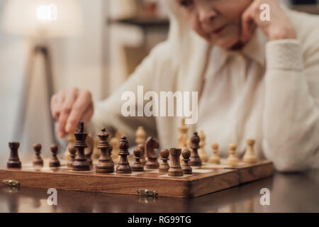 cropped view of senior woman sitting at table, propping chin with hand and playing chess at home Stock Photo