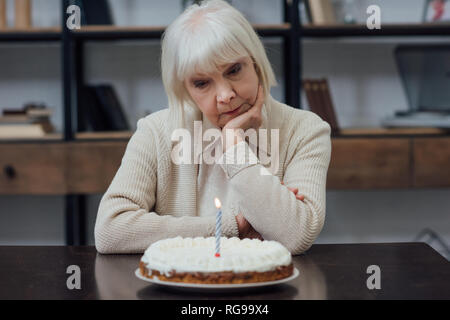 upset senior woman sitting at table, propping chin and looking at birthday cake with burning candle at home Stock Photo