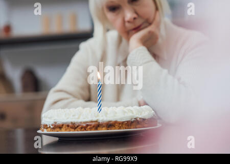 selective focus of sad senior woman sitting at table and looking at birthday cake with burning candle at home Stock Photo