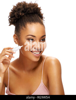 Pretty young girl with flawless skin using cream around the eyes. Photo of smiling african american girl on white background. Skin care and beauty con Stock Photo