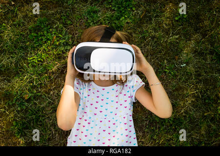Little girl wearing Virtual Reality Glasses lying on meadow in the garden Stock Photo