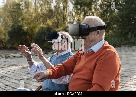 Two old friends sitting by the riverside, using VR glasses Stock Photo
