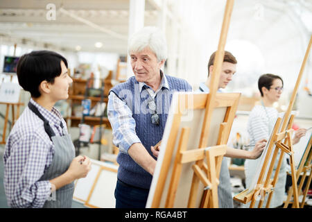 Portrait of mature art teacher painting picture on easel standing with group of students in art class, copy space Stock Photo