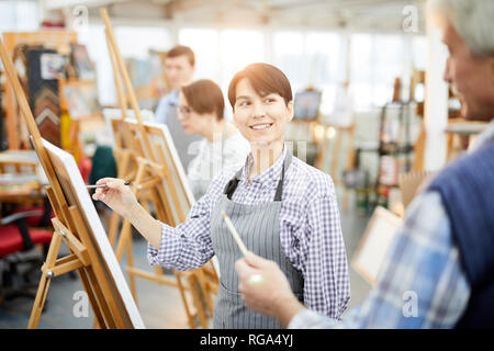 Waist up portrait of smiling young woman painting picture on easel in art class and looking at teacher, copy space Stock Photo