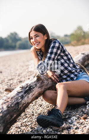 Happy young woman sitting at log on pebble shore Stock Photo