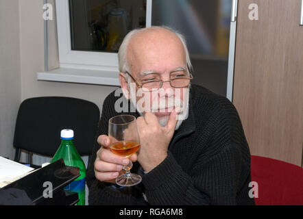 Close-up head and shoulders portrait of bald elderly man with grey long hair and beard in black knitted zipper sweater that is sitting with wine glass Stock Photo