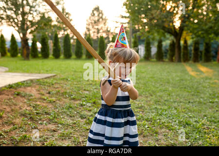 Happy little girl wearing party hat and holding wooden sword in garden Stock Photo