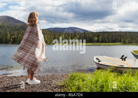 Finland, Lapland, woman wrapped in a blanket standing at the lakeside Stock Photo