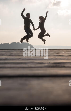 Happy young couple holding hands jumping in the air from a wooden pier. Stock Photo