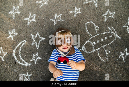 Portrait of smiling toddler wearing pilot hat and goggles lying on asphalt painted with airplane, moon and stars Stock Photo