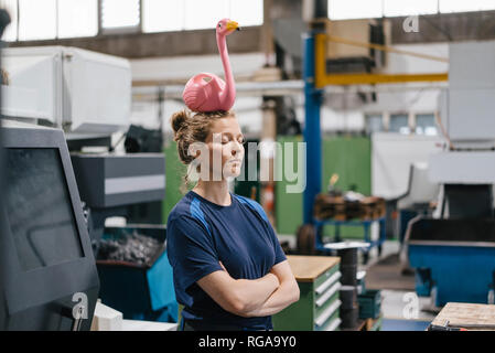 Young woman working as a skilled worker in a high tech company, balancing a pink flamingo on her head Stock Photo