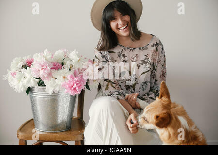 Stylish boho girl sitting at metal bucket with peonies on rustic wooden chair and playing with cute golden dog.Beautiful hipster woman playing with he Stock Photo