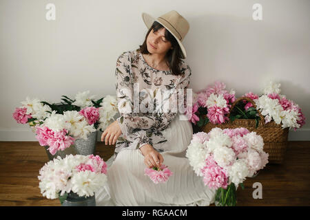 Boho girl sitting at pink and white peonies in rustic basket and metal bucket on wooden floor. Stylish hipster woman in bohemian dress holding pink pe Stock Photo
