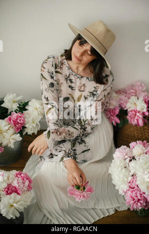 Boho girl sitting at pink and white peonies in rustic basket and metal bucket on wooden floor. Stylish hipster woman in bohemian dress with flowers. H Stock Photo