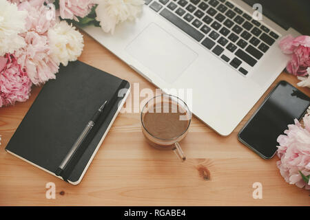 Stylish laptop, phone with empty screen, black notebook, coffee and peonies on rustic wooden table. Freelance concept. Girly workplace. Space for text Stock Photo