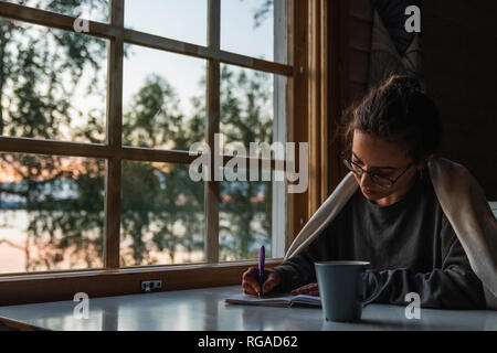 Finland, Lapland, young woman sitting at the window at a lake writing into diary Stock Photo