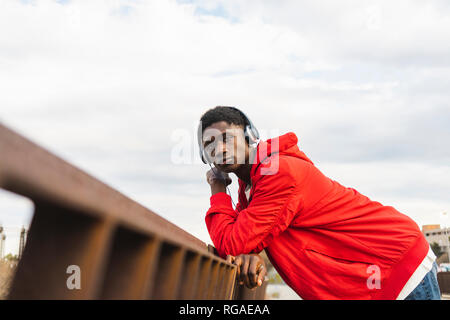 Young black man leaning on fence, listening music with headphones Stock Photo