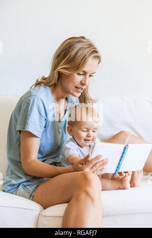 Mother and baby girl sitting on couch looking at picture book Stock Photo