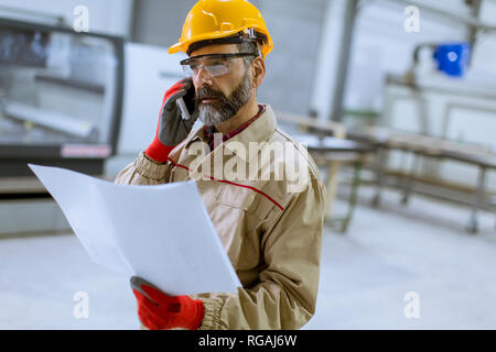 Handsome mature engineer looking at plan and using mobile phone in the factory Stock Photo