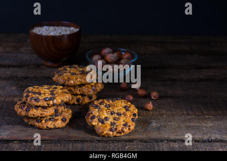 Avena cookies with chocolate chips and hazelnuts on a wood table with a bowl of avena flakes and another one of hazelnuts with shell Stock Photo
