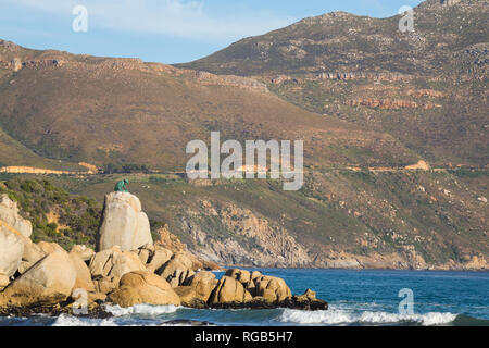 Chapmans Peak Drive along the mountain behind the bronze statue of the leopard on the rocks as seen from Hout Bay beach on a sunny Autumn afternoon Stock Photo