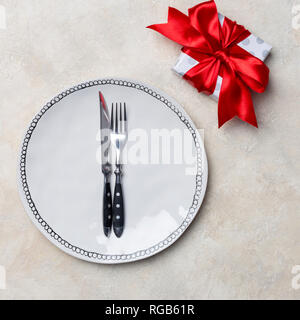 Holiday white plate with gift box with red ribbon, fork and knife near, at white background. Free space for text. Romantic decoration, square template Stock Photo