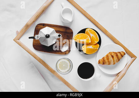 Romantic Valentines Day breakfast in bed, tray with fresh croissant, cup of coffee espresso with milk, glass of water with lemon and fresh oranges. Go Stock Photo